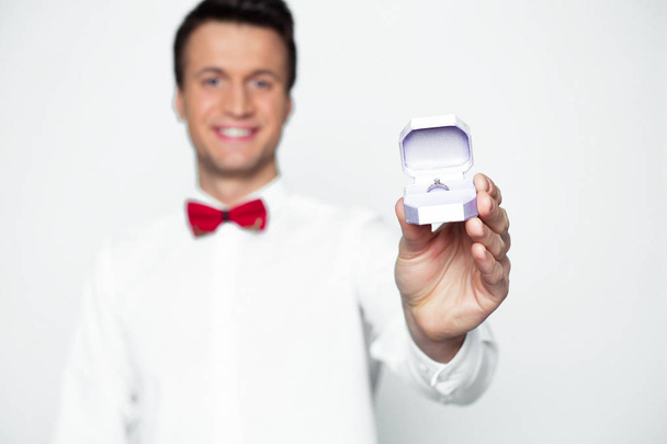 Make a proposal to your girlfriend. A handsome smiling modern elegant man in a white shirt and a red butterfly is holding a box with an engagement ring in front of the camera. - Foto, Imagen