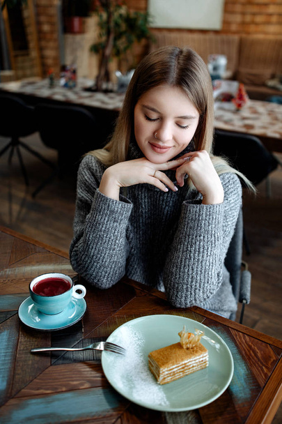 the girl the blonde in a warm winter sweater sits at a table in cafe and eats a dessert cake honey pie, the cup of tasty herbal tea or compote stands nearby. - Fotó, kép