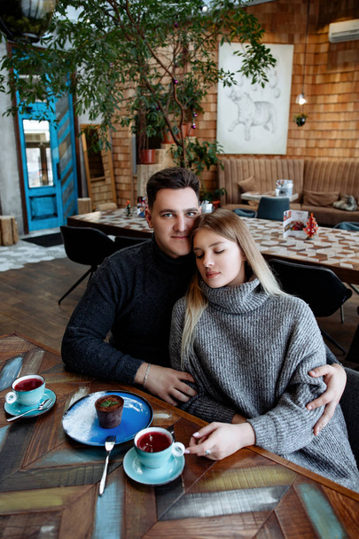 Young beautiful loving couple in cafe. The man and the woman in cozy gray sweaters communicate, embrace and smile. - Zdjęcie, obraz