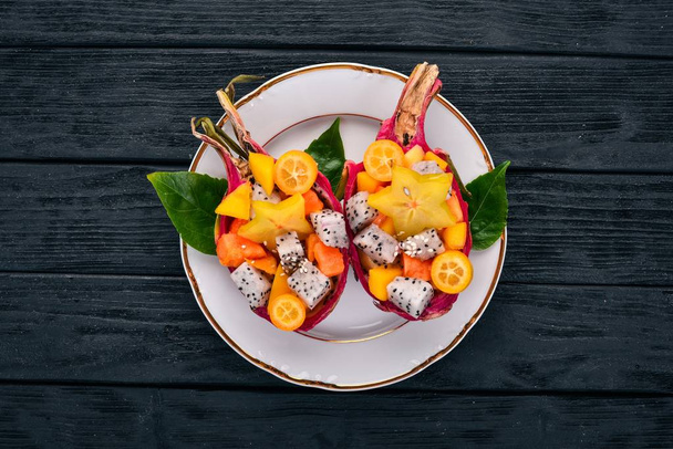 Tropisch Fruit salade geserveerd in de helft een Dragon Fruit. Papaya, ramboetan, tamarinde, cactus fruit, mango. Op een houten achtergrond. Bovenaanzicht. - Foto, afbeelding