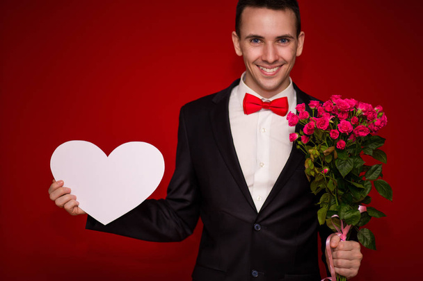 Πορτρέτο του νεαρός επιχειρηματίας γέλιο με λουλούδια και μεγάλο χαρτί καρδιά απομονώνονται σε κόκκινο φόντο - Φωτογραφία, εικόνα