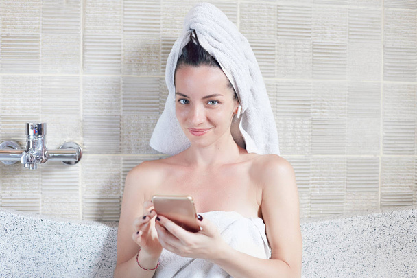 Mixed raced  young beautiful model with towel on her head sitting in bath tube and checking mail in modern waterproof mobile phone, speaking using bluetooth and wireless earphones. Technology concept. - Photo, Image