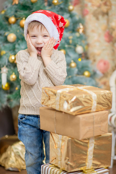 a boy in a red cap sits near a Christmas tree with a gift - Φωτογραφία, εικόνα