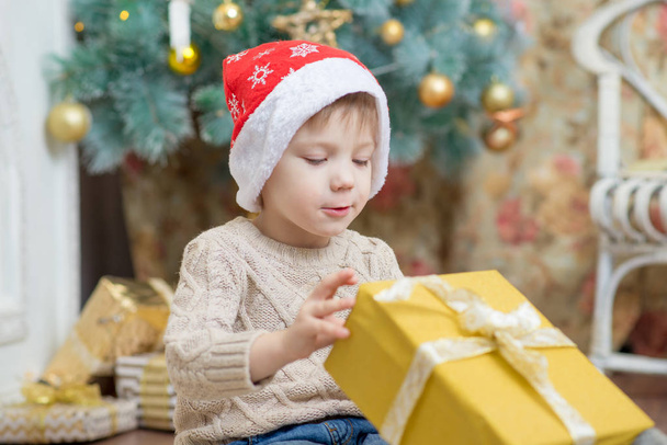 a boy in a red hat sits at a Christmas tree with a gift - Foto, Bild