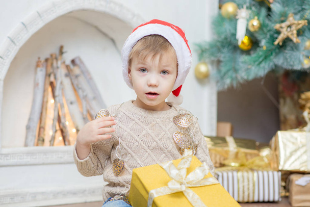 Kırmızı şapkalı bir çocuk bir Noel ağacı bir hediye ile oturuyor. - Fotoğraf, Görsel