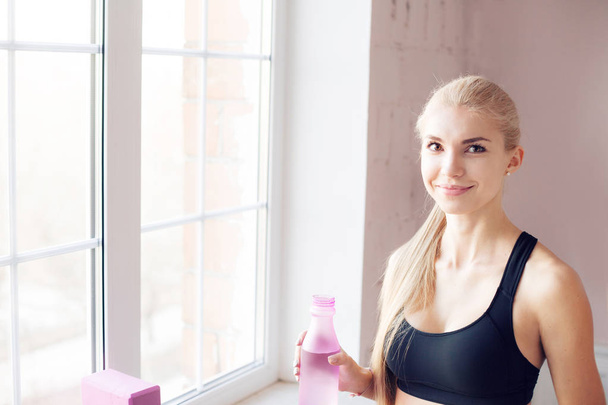home fitness. drink clean water from a bottle. Lovely young blonde girl. near the window - Фото, изображение