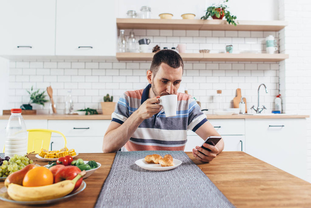 Молодой человек сидит за столом на кухне с чашкой кофе с помощью мобильного телефона
. - Фото, изображение