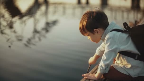 Schattig blanke jongen speelt met stok in de buurt van lake. Schattig kindje met rugzak op een pier. Gelukkige jeugd. 4k. - Video