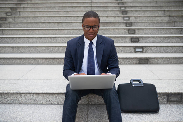 Alegre ejecutivo de negocios afroamericano sentado al aire libre y trabajando en el ordenador portátil
 - Foto, imagen