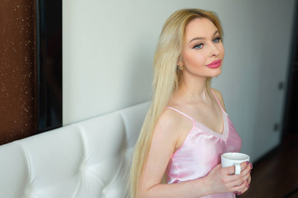 beautiful young blond woman with long hair in pink pajamas with a cup of drink in hands on a white bed in the bedroom - Фото, зображення