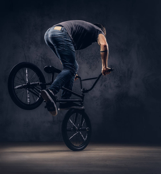 Handsome man in t-shirt and jeans makes an acrobatic figure with BMX in a studio. - Фото, зображення