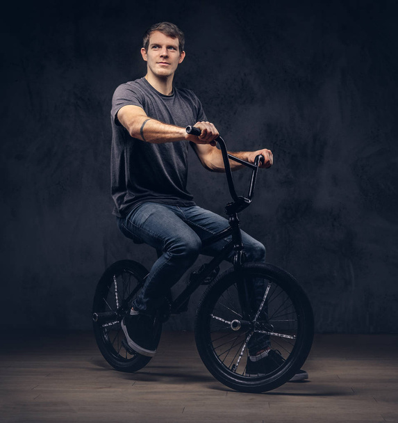 Handsome man in t-shirt and jeans sitting on a BMX. Isolated on a dark background. - Fotó, kép