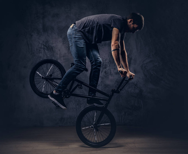 Handsome man in t-shirt and jeans makes an acrobatic figure with BMX in a studio. - Photo, Image