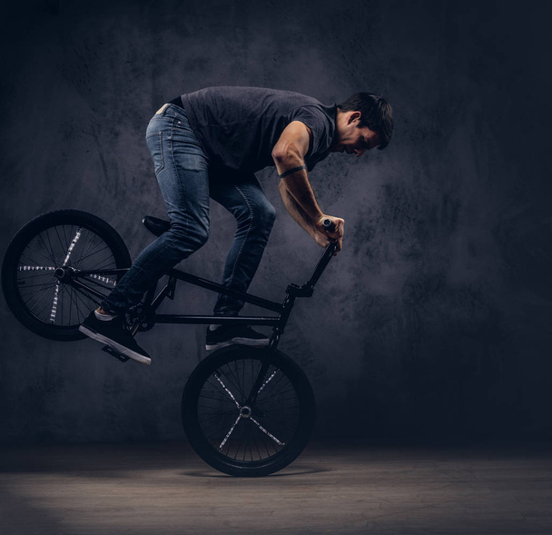 Handsome man in t-shirt and jeans makes an acrobatic figure with BMX in a studio. - Fotoğraf, Görsel
