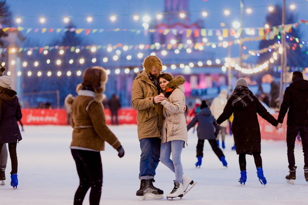 Ice skating rink and lovers together. A pair of young people in an embrace on a city skating rink lit by light bulbs and bright lights. Winter date for Christmas on the ice arena - Photo, Image