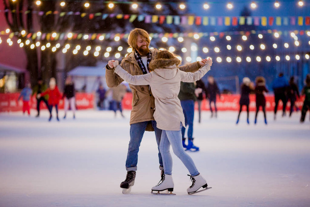Тематический каток и любящая пара. знакомство с молодыми, стильными людьми, катающимися рука об руку на городском катке, освещенном лампочками и огнями. Конькобежный спорт зимой на Рождество на ледовой арене - Фото, изображение