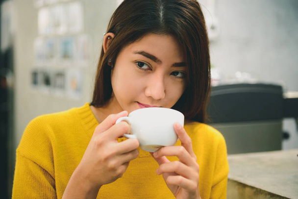 Eine lange asiatische junge Frau mit langen schwarzen Haaren, die ihren Kaffee am Wochenende bei guter Stimmung im Café hält und probiert. Verkostung Kaffee in modernen kleinen Laden. Konzepte der Lebensmittel- und Getränkeindustrie. - Foto, Bild