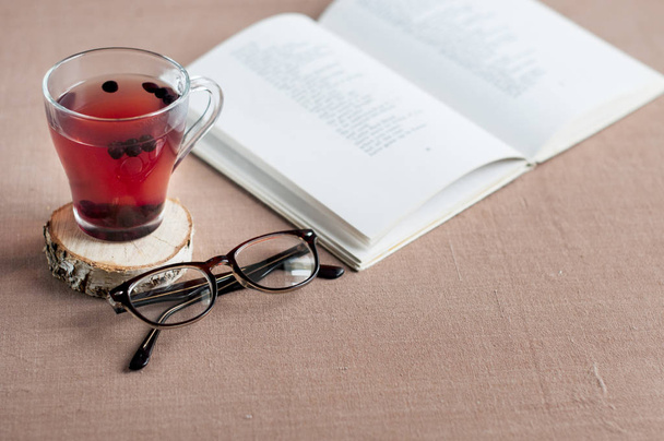 Blueberry tea in a clear glass cup on a round wooden stand with glasses and a open book on a brown tablecloth. - Photo, Image
