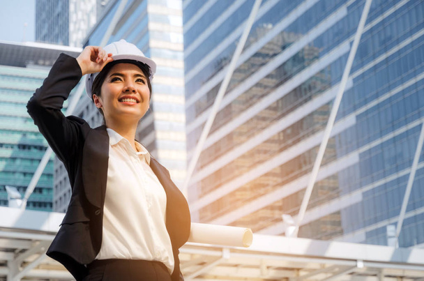 thinking business or engineer asian woman with white safety helmet in modern black suit in big city background, planning start up new project, successful, industry, architecture, construction concept - Foto, Imagen