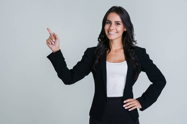 Take a look here! Beautiful young businesswoman pointing away and looking at camera with smile while standing against white background - Foto, imagen