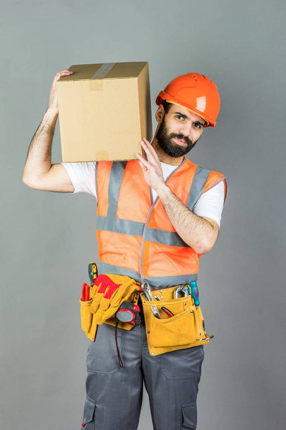 Άνδρας-οικοδόμος με ένα κράνος πορτοκαλί με ένα κουτί από χαρτόνι με τα χέρια του. Σε γκρι φόντο. - Φωτογραφία, εικόνα