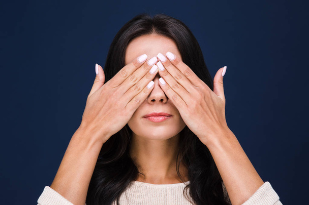 Verstecken. Porträt einer schönen jungen Frau, die ihre Augen mit den Händen bedeckt, während sie vor schwarzem Hintergrund steht - Foto, Bild