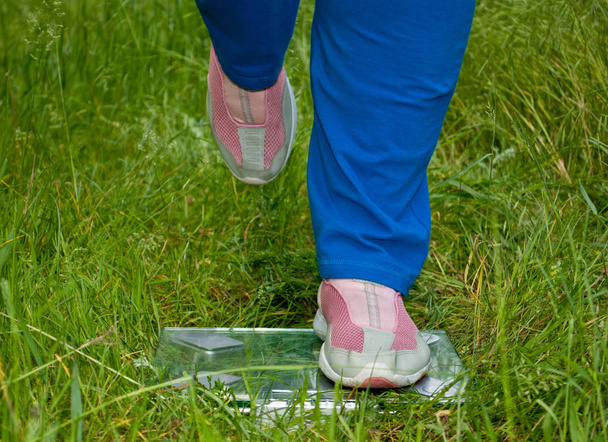 Sport atleet dikke vrouw verliezen gewicht staande op schalen benen linkerbeen verhoogd blauw sport broeken knieën in het roze sneakers glas transparant schubben op groen gras wazig achtergrond, vooraanzicht - Foto, afbeelding