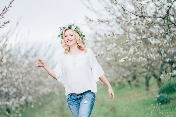 Hermosa joven mujer embarazada alegre en corona de flores en la cabeza tocando el vientre mientras camina y baila en el jardín de árboles de primavera. Beauty People Conceptos de estilo de vida. Feliz mamá en espera de bebé
! - Foto, Imagen