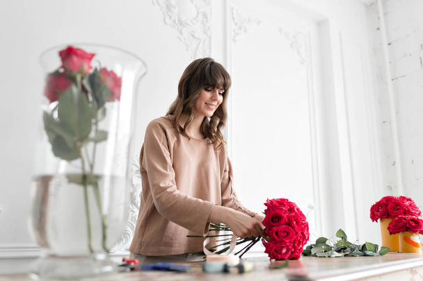 Усміхаючись мила молода жінка-флорист, організовує рослини в квітковому магазині. Люди, бізнес, продаж та концепція флористики. Букет червоних троянд
 - Фото, зображення