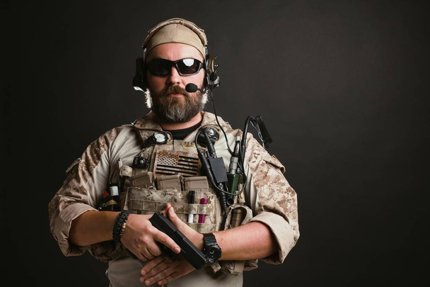 Un homme brutal en uniforme militaire et gilet pare-balles se tient debout et tient un pistolet sur un fond noir dans le studio. Le joueur barbu dans les lunettes de sécurité airsoft ou lunettes et écouteurs actifs
 - Photo, image