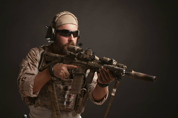 Brutaler Mann in militärischer Wüstenuniform und Rüstung steht in einem Gefechtsstand und hält sein Gewehr vor schwarzem Hintergrund. der bärtige Spieler mit der Airsoft-Schutzbrille, der auf ein Gewehr zielt - Foto, Bild