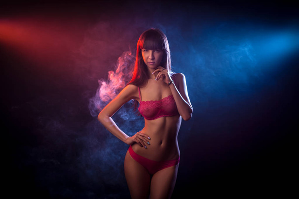 Young dark-haired woman in red lingerie posing against a background of red and red smoke from a wipe on a black isolated background - 写真・画像
