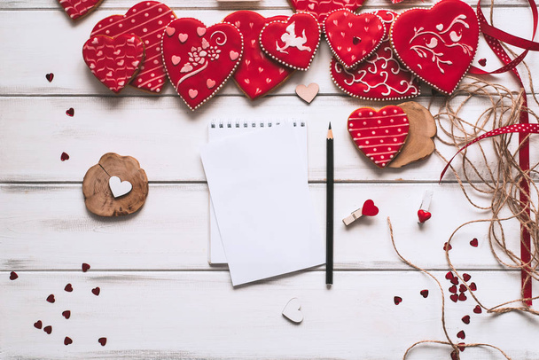 Composición festiva con galletas acristaladas rojas, papel y lápiz para letras románticas sobre tablones de madera de fondo para el día de San Valentín
. - Foto, imagen