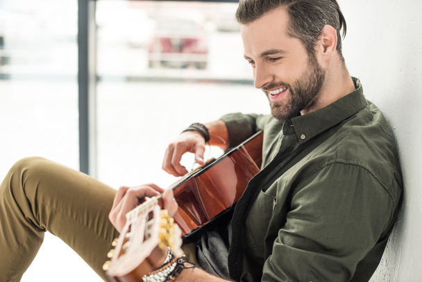 boční pohled hezký usmívajícího se muže hrál na akustickou kytaru - Fotografie, Obrázek