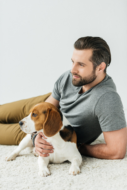 schöner Mann liegt mit niedlichem Beagle auf Teppich und schaut weg - Foto, Bild