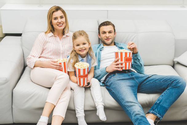 smiling family with popcorn watching film together at home - 写真・画像