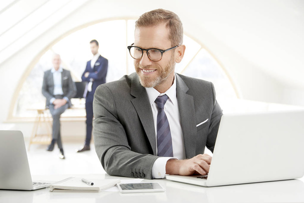 Middle aged businessman wearing suit and smiling while sitting at office and working on laptop. - Foto, Imagem