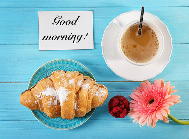 Plate with croissant, cup of coffee and flower on table, top view. Tasty breakfast and "Good morning" wish - Photo, Image
