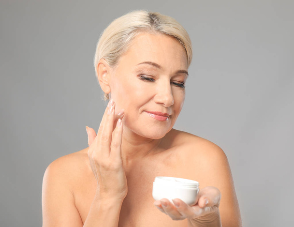 Portrait of beautiful mature woman with jar of cream on grey background. Skin care concept - 写真・画像