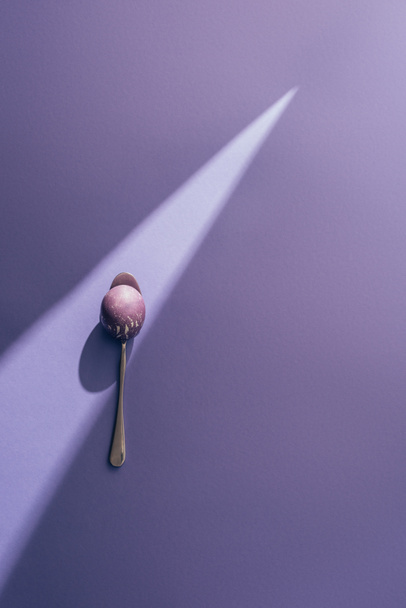 Пасхальное окрашенное яйцо в ложку на фиолетовом фоне с солнечным лучом
 - Фото, изображение