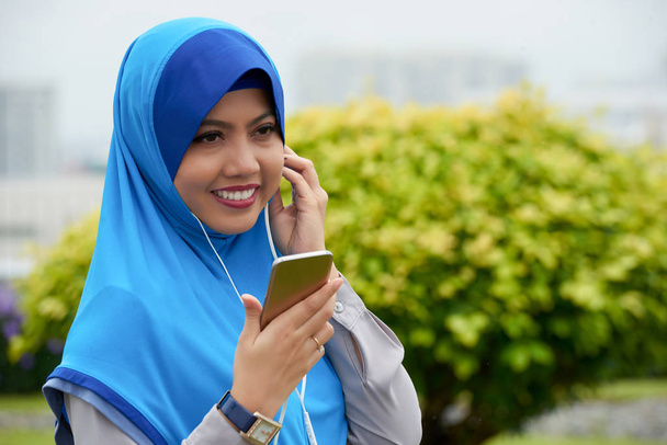 Portret van jonge aantrekkelijke vrouw in hijab bedrijf smartphone, luisteren naar muziek en lachende - Foto, afbeelding