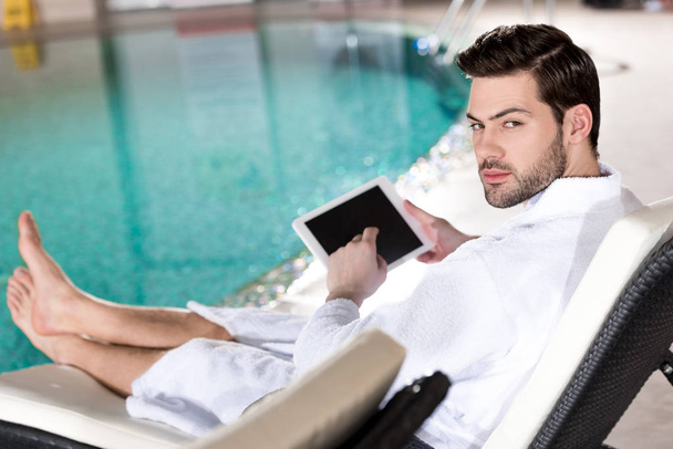handsome man in bathrobe using digital tablet and looking at camera in spa center - Fotó, kép
