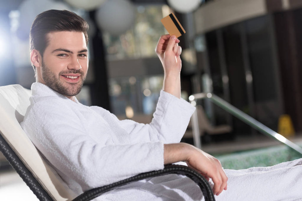 young man in bathrobe holding credit card and smiling at camera in spa center    - Foto, Bild