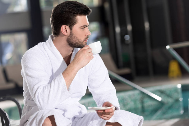 handsome young man in bathrobe drinking coffee and looking away in spa center - Fotografie, Obrázek