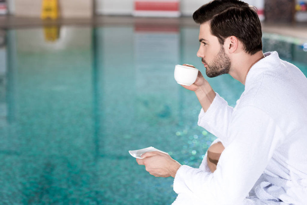 side view of man in bathrobe drinking coffee while sitting near pool in spa center - Foto, imagen