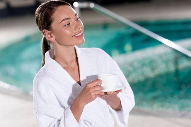 beautiful young woman in bathrobe holding cup of coffee near pool - Foto, afbeelding