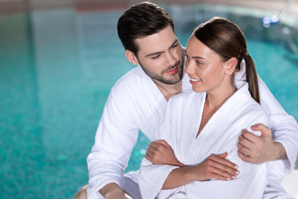 feliz jovem casal em roupões de banho abraçando perto da piscina no centro de spa
 - Foto, Imagem