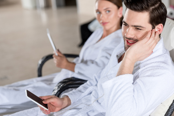 junges Paar in Bademänteln mit digitalen Geräten beim gemeinsamen Ausruhen im Wellness-Center - Foto, Bild