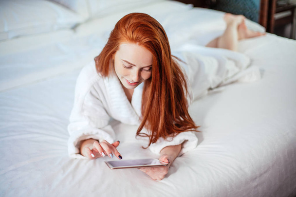 beautiful woman wearing bathrobe and using digital tablet while relaxing on bed - Photo, Image