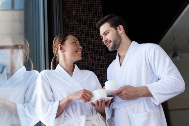 couple in bathrobes holding cups of tea and smiling each other in spa center - Φωτογραφία, εικόνα